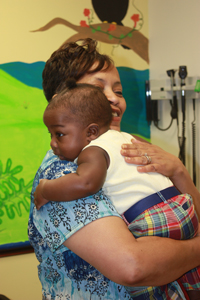 A smiling nurse in blue scrubs and yellow flowers is holding and hugging a toddler.